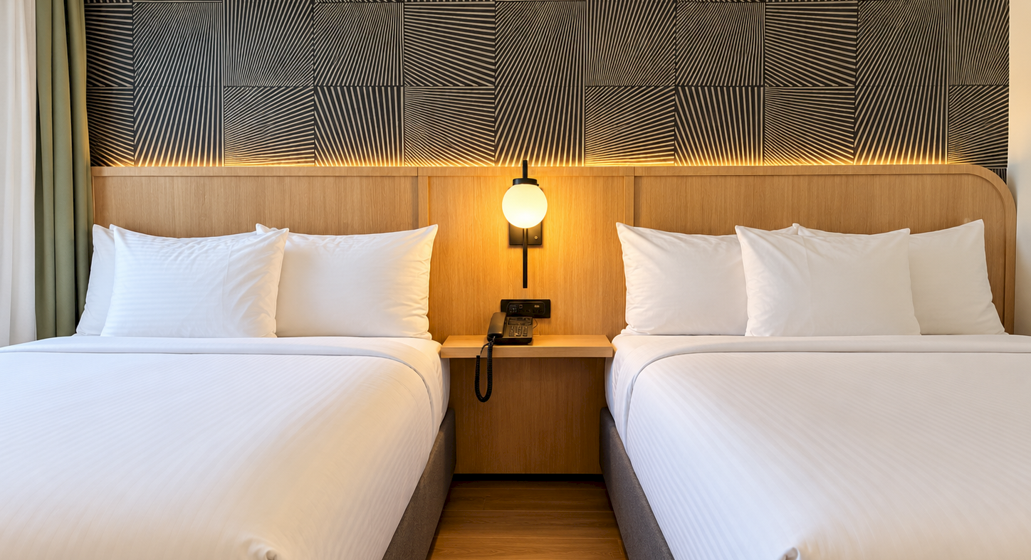 A modern hotel room with two neatly made beds, a wooden headboard, bedside phone, and warm wall lamp above.