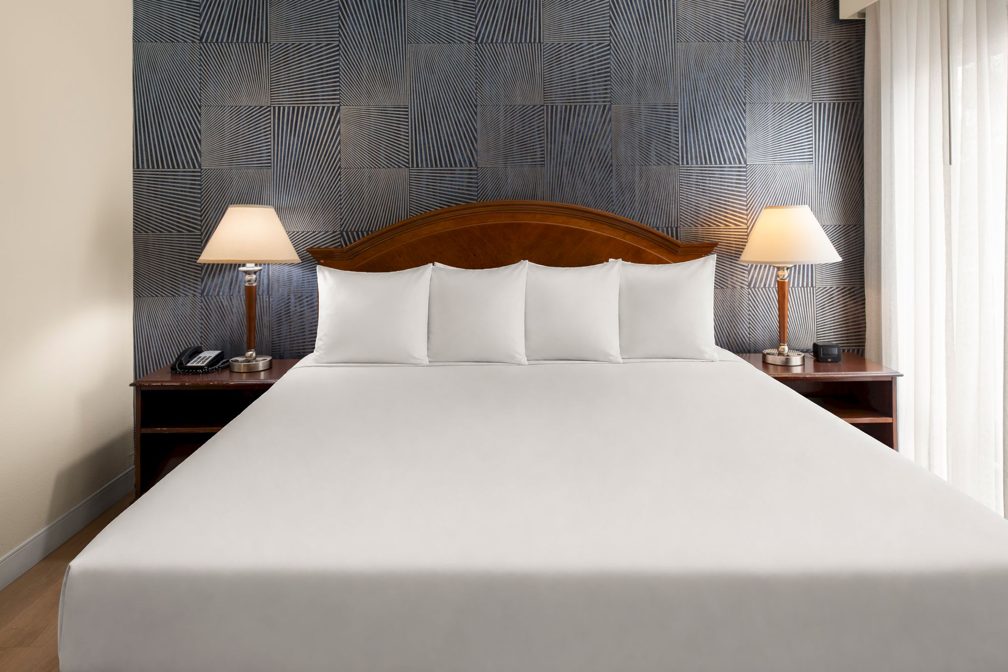 A neatly made hotel bed with white linens, two pillows, wooden headboard, two matching bedside tables, lamps, and a textured accent wall.