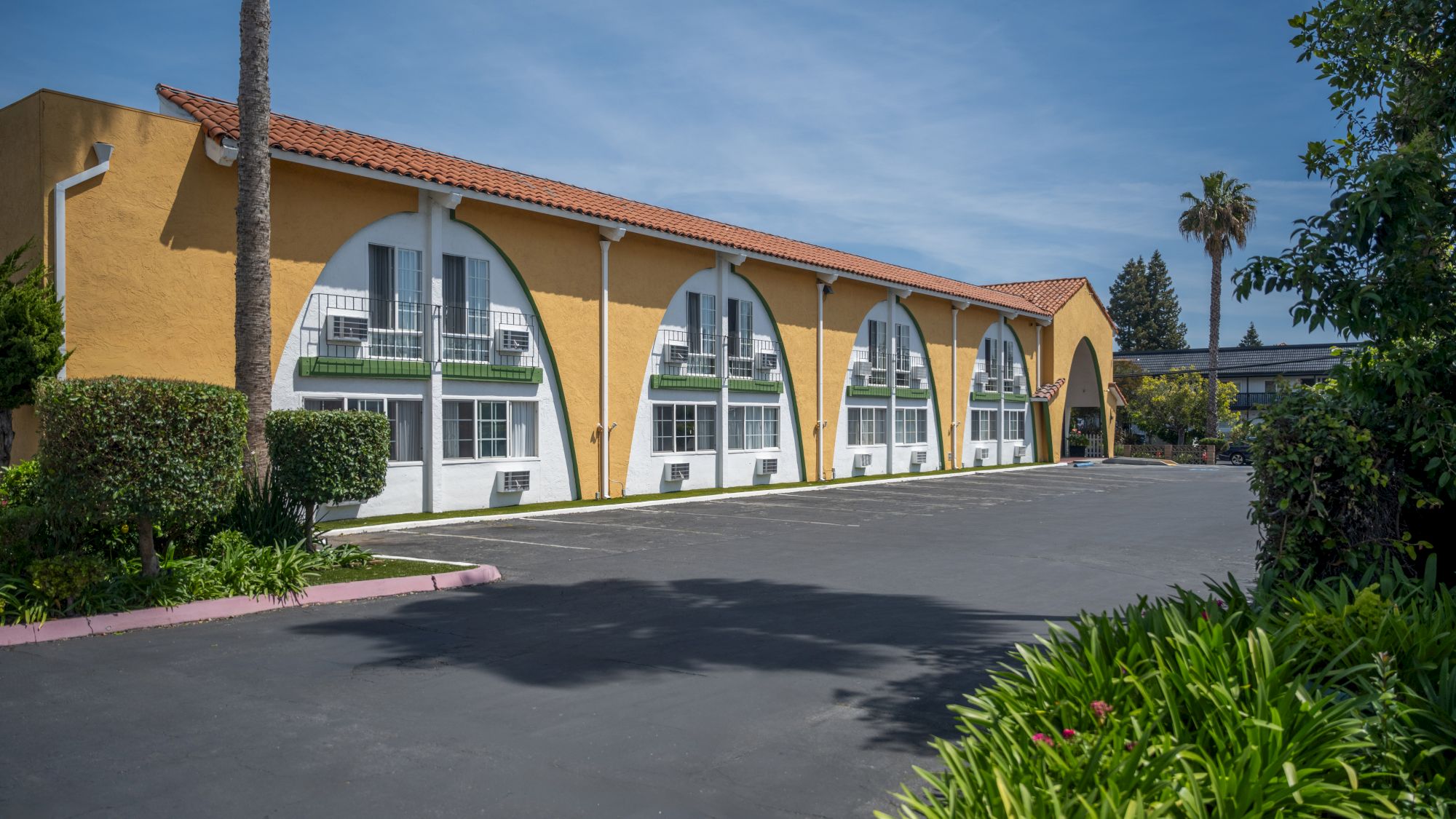 A row of yellow storefronts with arched windows and doors, a paved lot, palm trees, and tidy landscaping under a clear blue sky.