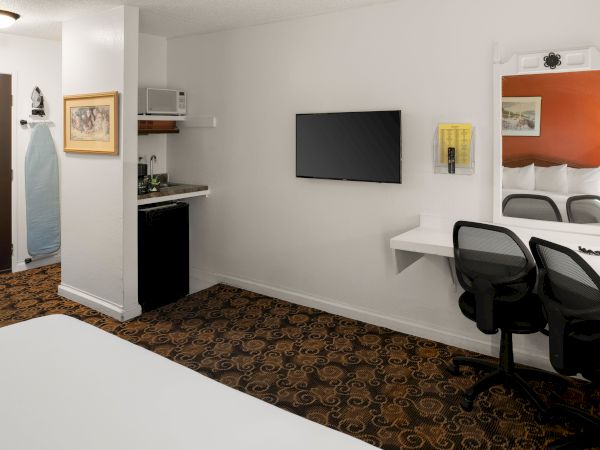 A small hotel room with a bed, a wall-mounted TV, a desk and chairs, a mirror, and a door leading to a compact kitchenette area.