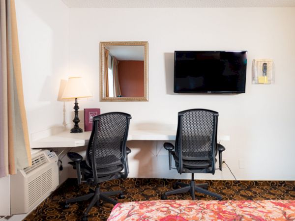 A hotel room desk area with two chairs, a wall‑mounted TV, a mirror, a lamp, and a window drape beside an air conditioner.