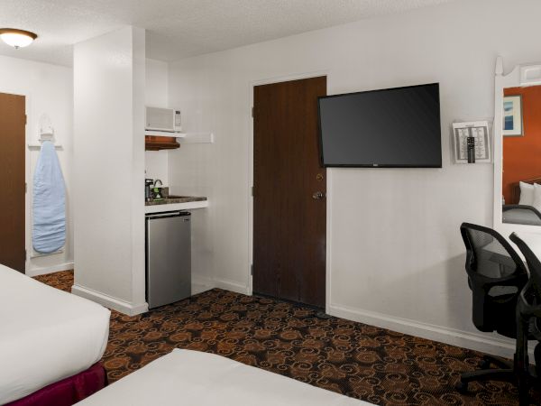 A hotel suite showing a bed, small kitchenette, wall-mounted TV, dark door, desk chair, and a doorway to a bath/upstairs area. End.