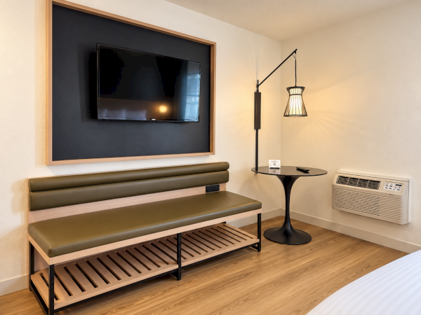 A modern room corner with a wall-mounted TV, green bench seating, a side table, hanging lamp, and an air conditioner/heater.