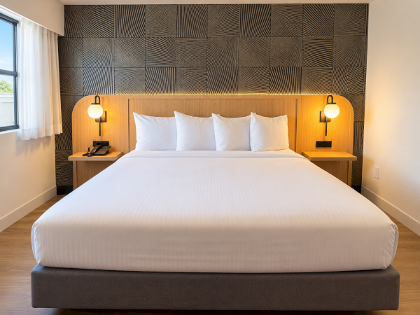 A modern hotel room with a large white bed, wooden headboard, side tables, and warm wall lamps.