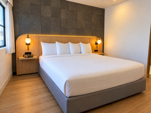 A modern hotel room with a large bed, white pillows and fitted lamps against a geometric wall backdrop, lit warmly.