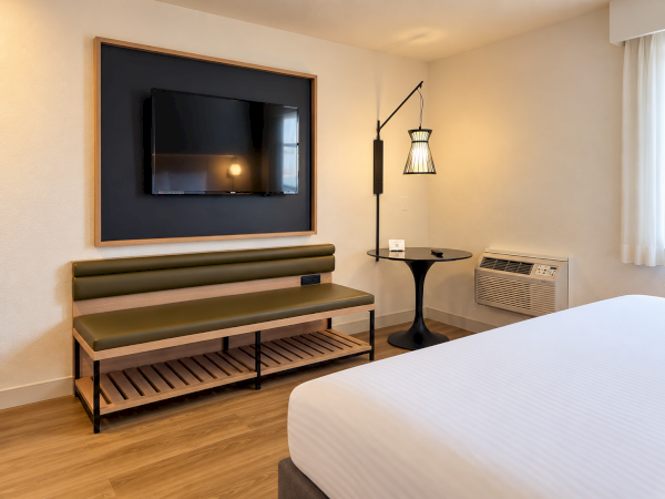 A modern bedroom with a wall-mounted TV, cushioned bench, pendant lamp, side table, and an AC unit near a window.