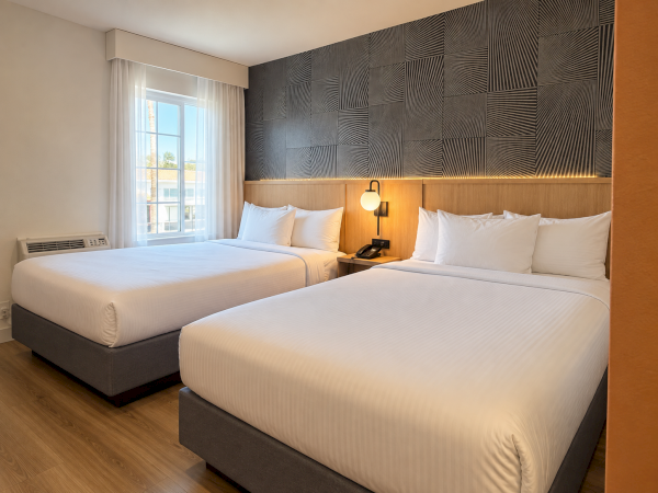 A modern hotel room with two neatly made beds, white linens, wood headboards, geometric wall panels, and a bedside lamp.