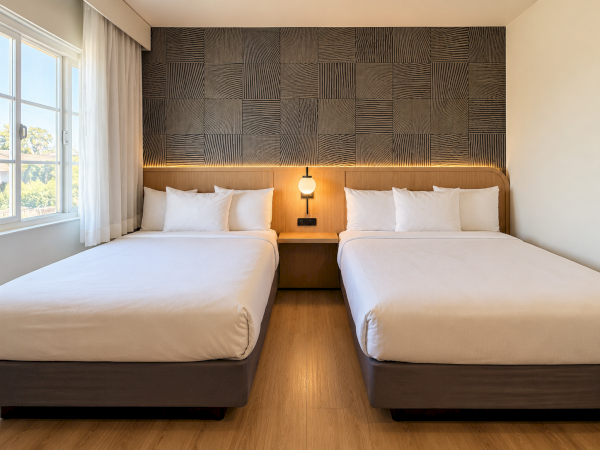 A modern hotel room with two twin beds, a wood-paneled headboard, bedside lamp, and large window with sheer curtains.