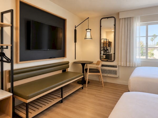 A modern hotel room with a large wall TV, leather bench seating, round table and chair, pendant lamp, and bright window view.