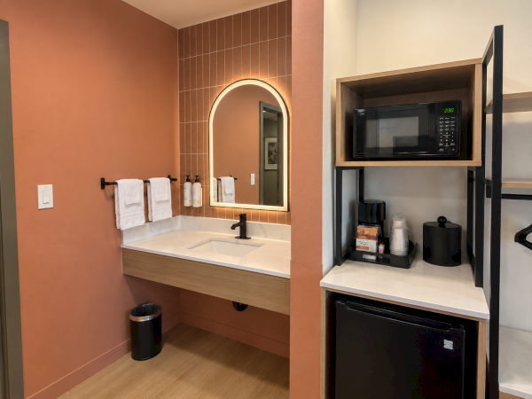 A modern bathroom vanity with a lit mirror, towels, and a built-in kitchenette area with a microwave, coffee maker, and mini fridge.