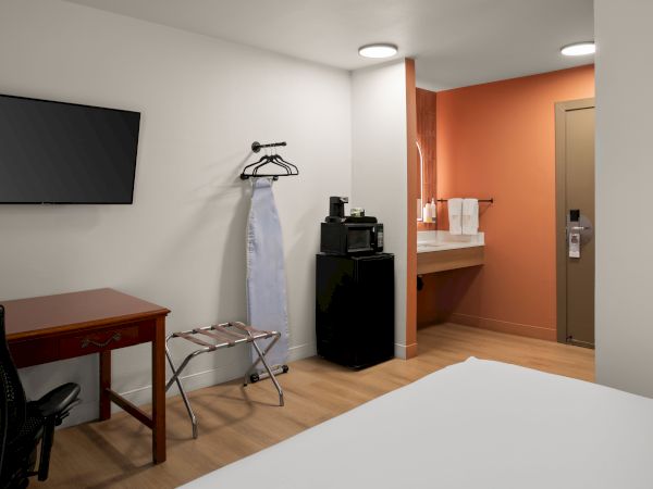 A hotel room with a desk and chair, wall-mounted TV, ironing board, minibar, and a small kitchenette/vanity area near an orange accent wall.