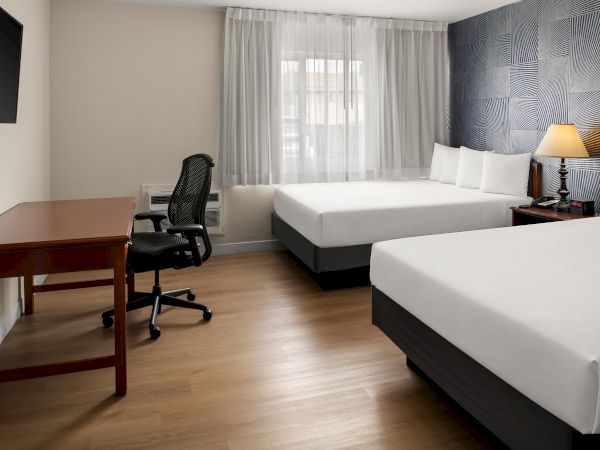 A hotel room with two single beds, a desk and chair, a wall-mounted TV, a window with sheer curtains, and a nightstand with a lamp.