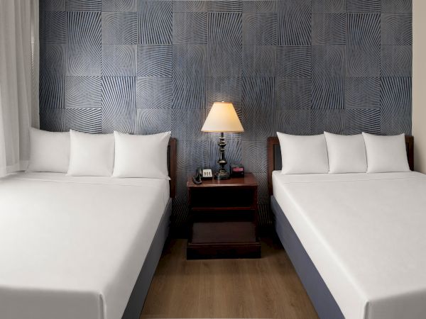 Two single beds with white linens face each other, a shared nightstand with a lamp between them, and a patterned blue headboard wall.