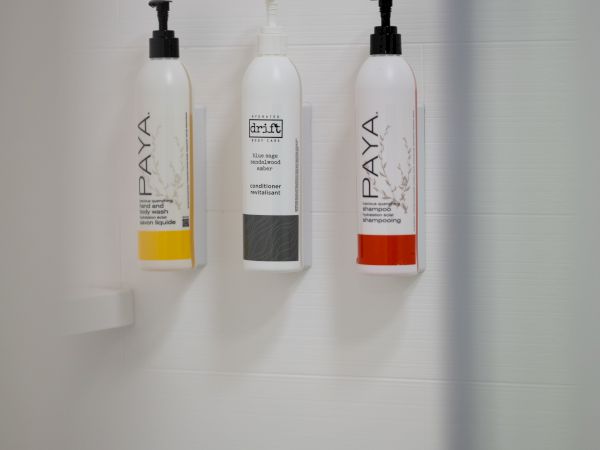 A bathroom shower with three wall-mounted bottles (two white with yellow and red accents and one gray label) in a minimalist setup, seen through a glass door.