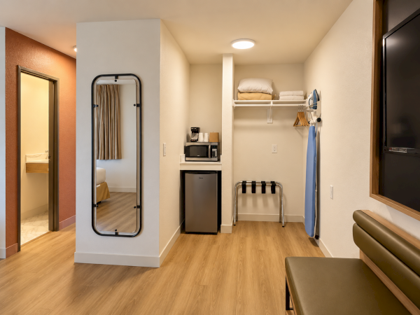 A modern hotel room corner with a full-length mirror, coffee maker and microwave, mini fridge, ironing board, and folded towels.