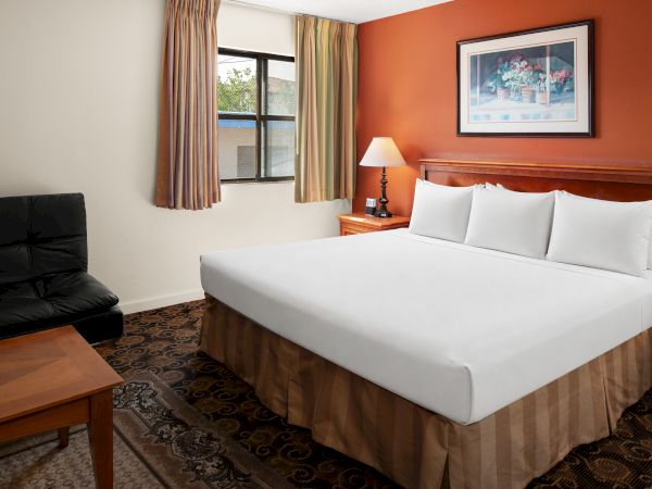 A hotel room with a king bed, two nightstands, lamps, a sofa, and a window with beige curtains. (140 chars max, ending with a period)