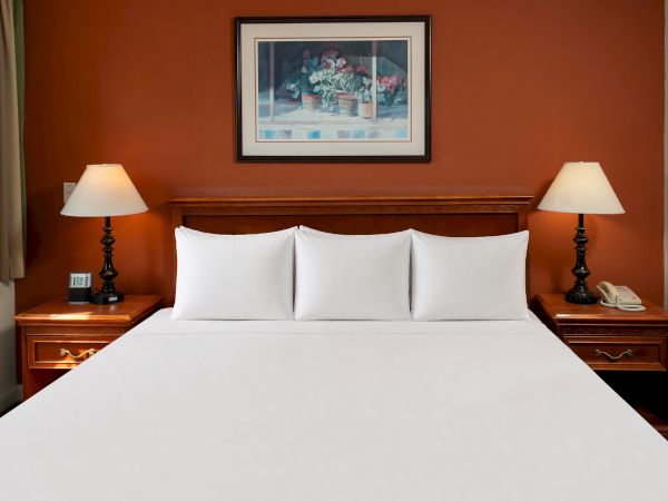 A tidy hotel room with a large bed, white linens, two nightstands, matching lamps, and a framed artwork above the headboard.