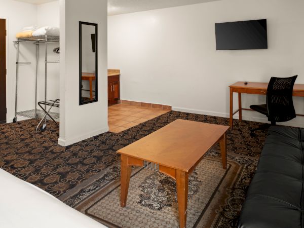 A small living area with a patterned rug, wooden coffee table, a black sofa, a wall-mounted TV, a wooden desk with chair, and a coat rack near a doorway.