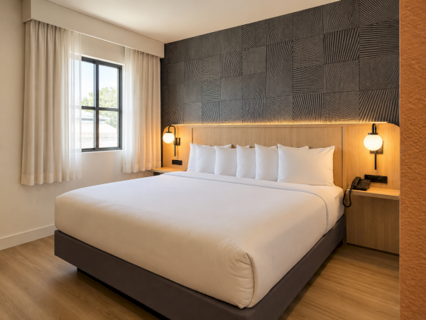 A modern hotel bedroom with a large bed, white pillows and duvet, bedside lamps, and a geometric wall with a window.
