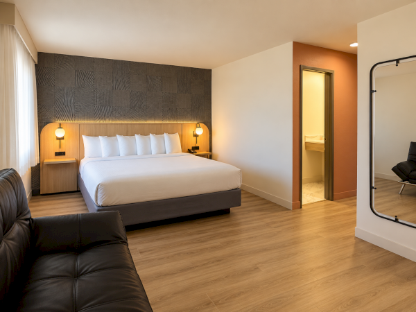 A modern hotel bedroom with a neatly made bed, warm wall lighting, patterned headboard, and an open bathroom doorway.