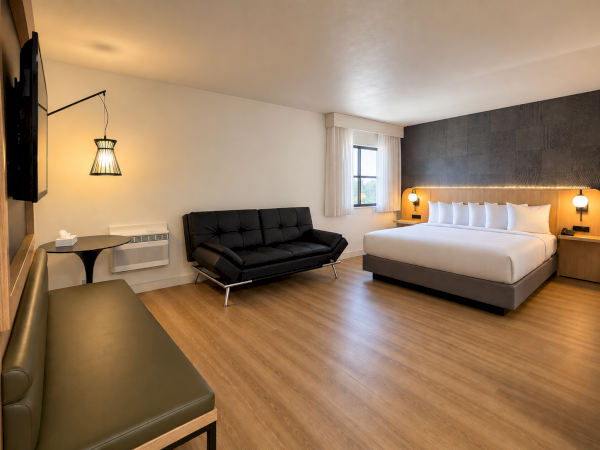 A modern hotel room with a black sofa, bedside lamps, a large bed, and wood floors.