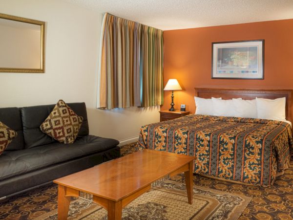 A cozy hotel room with a king bed, nightstands and lamps, a dark sofa with patterned cushions, a wooden coffee table, and a warm orange accent wall.