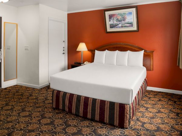 A hotel room with a bed against a red accent wall, two lamps, a framed painting, a patterned carpet, and a doorway with a full-length mirror nearby, ending with a period.