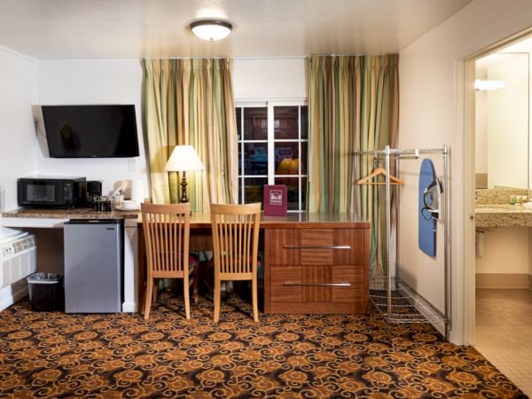 A hotel room with a desk, two chairs, a small fridge, microwave, TV, coat rack, and a doorway to a bathroom, all under patterned carpet.