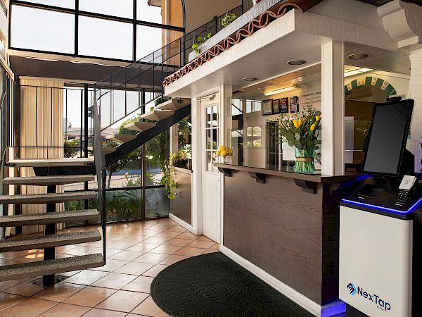 A modern hotel lobby with a reception desk, staircase to a mezzanine, large windows, potted plants, and a self-check-in kiosk nearby.