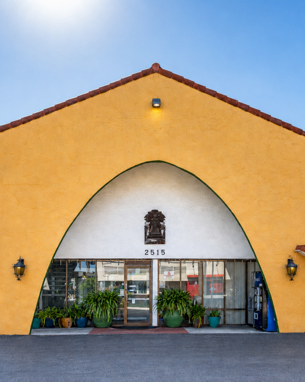 A yellow, arch-front building with a curved white entrance, black sign &ldquo;GRANADA&rdquo; nearby, potted plants, and a bright blue sky behind.