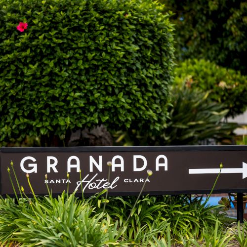 A hotel sign reads &ldquo;Granada&rdquo; with an arrow, set in a lush garden with manicured shrubs and colorful flowers nearby.
