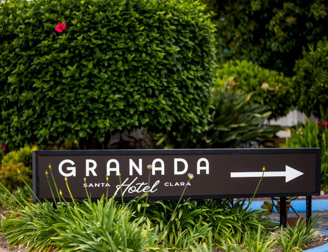 A hotel sign reads &ldquo;Granada&rdquo; with an arrow, set in a lush garden with manicured shrubs and colorful flowers nearby.