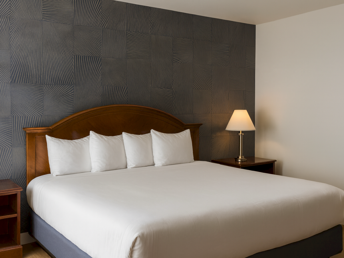 A neatly made hotel bed with white linens and four pillows, dark wood headboard, patterned accent wall, and a bedside table with a lamp.