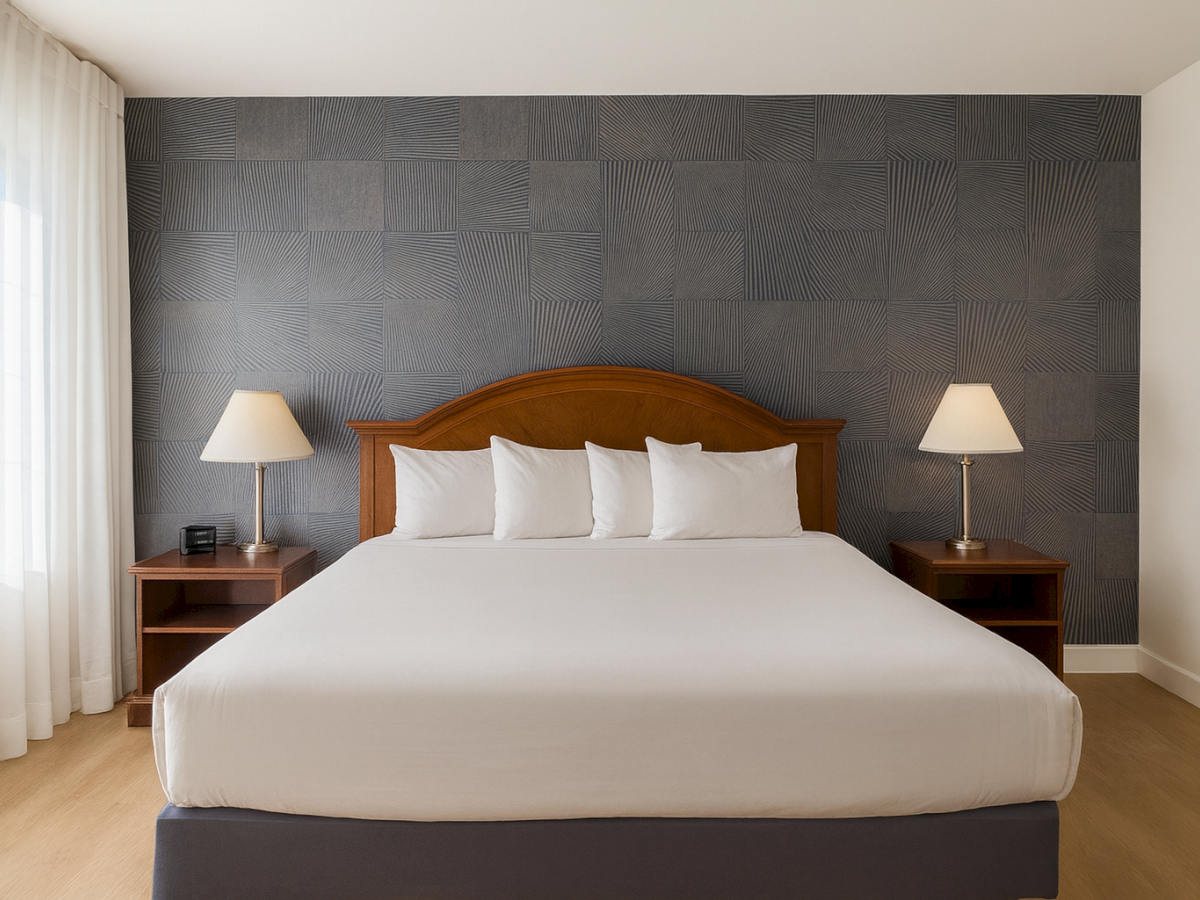A neatly made hotel bed with white linens and five pillows, dark wood headboard, two matching nightstands, lamps, and a textured dark accent wall.