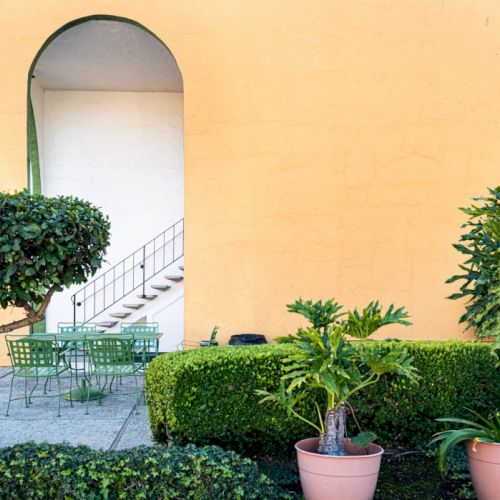 A sunny courtyard with a small blue pool, potted trees and flowers, an arched doorway, and warm orange walls.