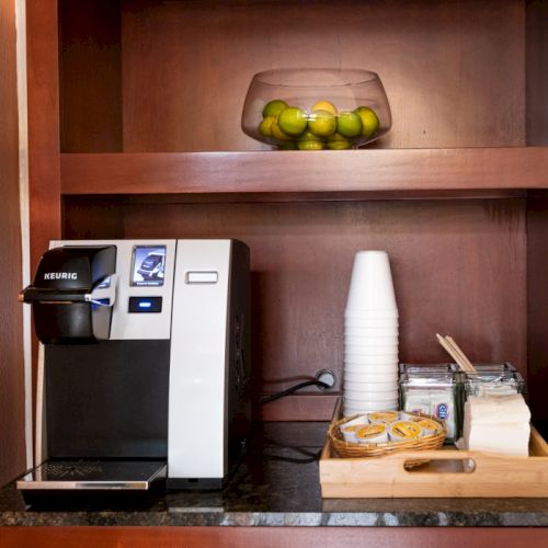 A small kitchen shelf with a black coffee maker, a stainless kettle, a jar, and a few containers, all on a dark wooden wall shelf.