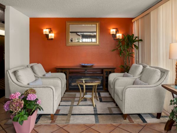 A cozy hotel lobby with two beige armchairs facing a wooden console, orange accent wall, glass coffee table, and plants, warmly lit.