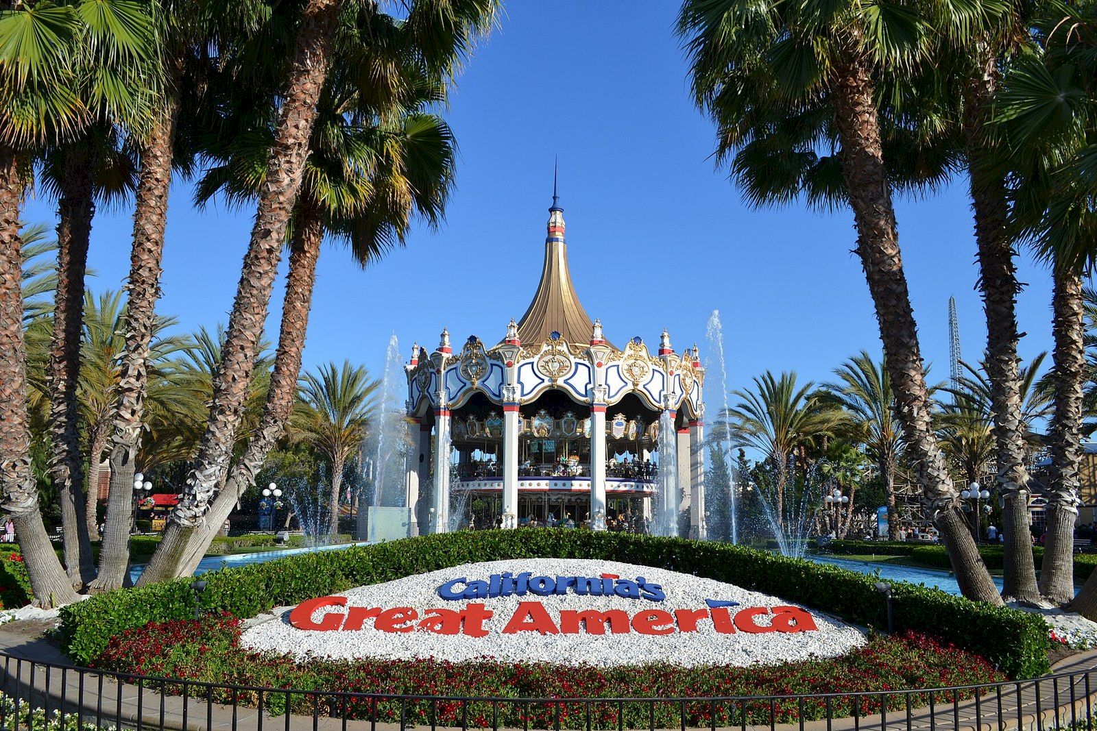 Palm trees frame a blue-and-white carousel at California&rsquo;s Great America, with a sunlit sky and fountain sprays.