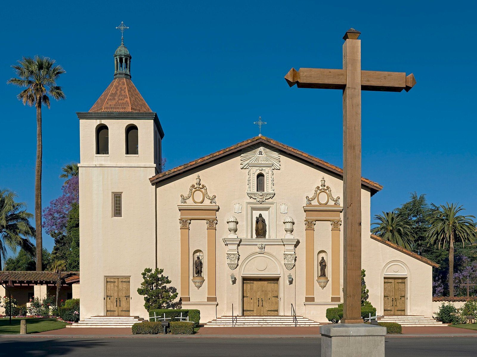 A white Spanish-style church fa&ccedil;ade with a large wooden cross, bell tower, and palm trees under a clear blue sky.