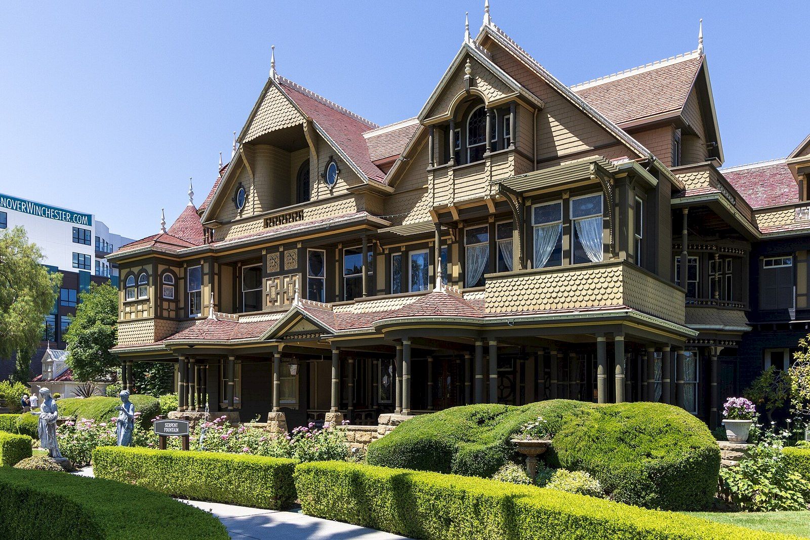 An ornate historic house with gabled roof, columns, and manicured hedges in a sunny garden courtyard.