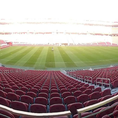 View from inside a football stadium with red seats and a green pitch, under bright daylight.
