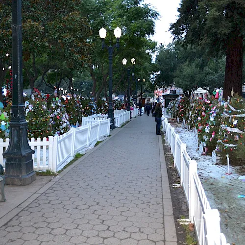 A festive holiday walkway with lit trees, colorful decorations, white fencing, and people strolling along a paved path.