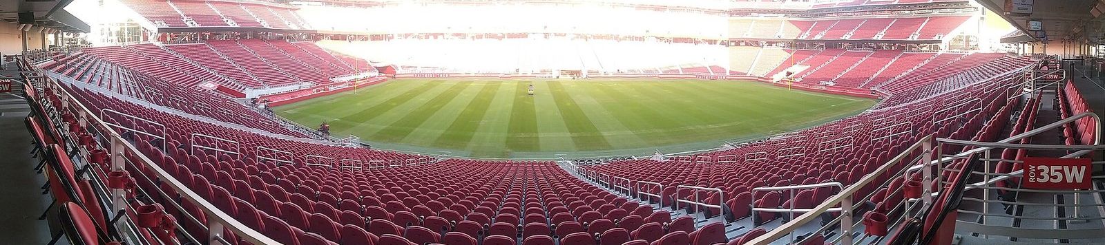 A wide view of an empty red-seated stadium with the green soccer field in the center, bright daylight outside the stands.
